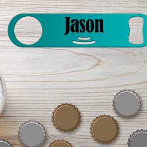 Puede incluir: Abrebotellas de metal azul turquesa con una inscripción negra que dice "Jason". El abrebotellas está sobre una superficie de madera clara con varias tapas de botellas esparcidas alrededor.