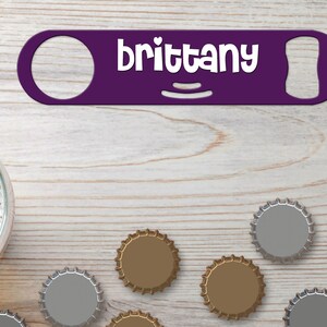 Puede incluir: Abrebotellas morado con el nombre "brittany" en letras blancas. El abrebotellas está sobre una superficie de madera clara con varias tapas de botellas esparcidas a su alrededor.
