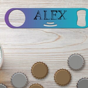 Puede incluir: Un abridor de botellas de metal azul y morado con el nombre "ALEX" impreso en él. El abridor está sobre una superficie de madera con varias tapas de botellas esparcidas a su alrededor.