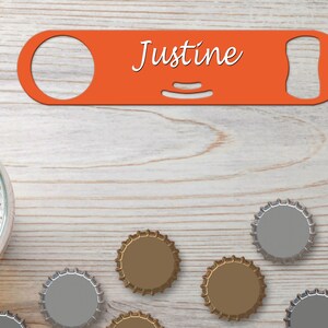 Puede incluir: Un abridor de botellas rojo con el nombre "Justine" impreso en él. El abridor está sobre una superficie de madera con varias tapas de botellas esparcidas a su alrededor.