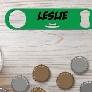 Puede incluir: Abrebotellas de metal verde con el nombre "LESLIE" impreso en letras negras. El abrebotellas está sobre una superficie de madera clara con varias tapas de botellas esparcidas a su alrededor.