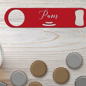 Puede incluir: Abrebotellas de metal rojo con el nombre "Pam" grabado en él. El abrebotellas está sobre una superficie de madera clara con varias tapas de botellas esparcidas a su alrededor.
