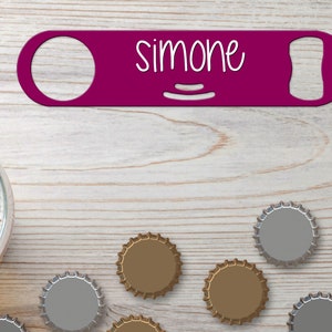Puede incluir: Un abridor de botellas morado con el nombre "Simone" escrito en él. El abridor de botellas está sobre una superficie de madera con varias tapas de botellas esparcidas a su alrededor.