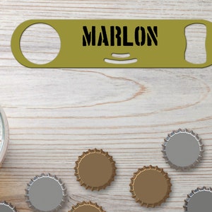 Puede incluir: Un abridor de botellas de metal dorado con el nombre "MARLON" grabado en él. El abridor de botellas está sobre una superficie de madera con varias tapas de botellas.