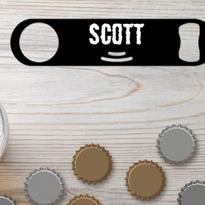 Puede incluir: Abrebotellas de metal negro con el nombre "SCOTT" impreso en blanco. El abrebotellas está sobre una superficie de madera clara con varias tapas de botellas esparcidas a su alrededor.