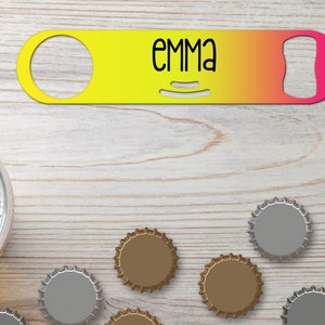 Puede incluir: Un abridor de botellas amarillo y rosa con el nombre "EMMA" impreso en él. El abridor está sobre una superficie de madera con varias tapas de botellas esparcidas a su alrededor.