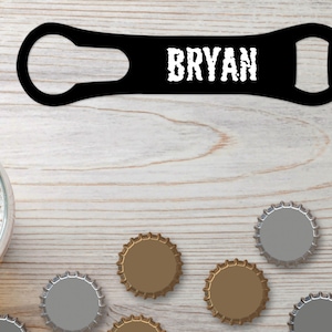 May include: Black bottle opener with the name "BRYAN" printed in white. The opener is on a light wood surface with several bottle caps scattered around it.