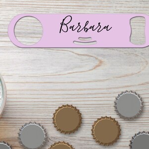 Puede incluir: Un abrebotellas rosa claro con el nombre "Barbara" en escritura negra. El abridor está sobre una superficie de madera clara, con tapas de cerveza y un vaso de cerveza en el fondo.