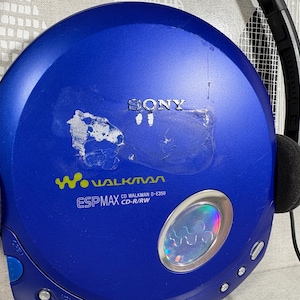 May include: A blue Sony Walkman CD player with black headphones attached. The device features the text SONY, Walkman, ESP MAX, and CD Walkman D-E350 CD-R/RW. There is a circular silver accent with a holographic center and visible adhesive residue.