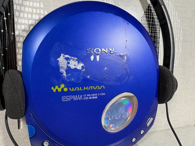 May include: A blue Sony Walkman CD player with black headphones attached. The device features the text SONY, Walkman, ESP MAX, and CD Walkman D-E350 CD-R/RW. There is a circular silver accent with a holographic center and visible adhesive residue.