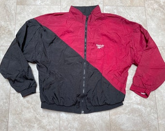 Puede incluir: Una chaqueta cortavientos negra y burdeos con cierre de cremallera y un logotipo de Reebok en el pecho derecho.