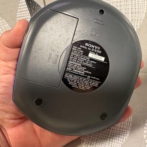 May include: A dark gray Sony portable CD player, model D-E350, featuring a circular design with a battery compartment door labeled OPEN. The back panel includes a black circular sticker with technical specifications, serial number 5732082, and regulatory text.