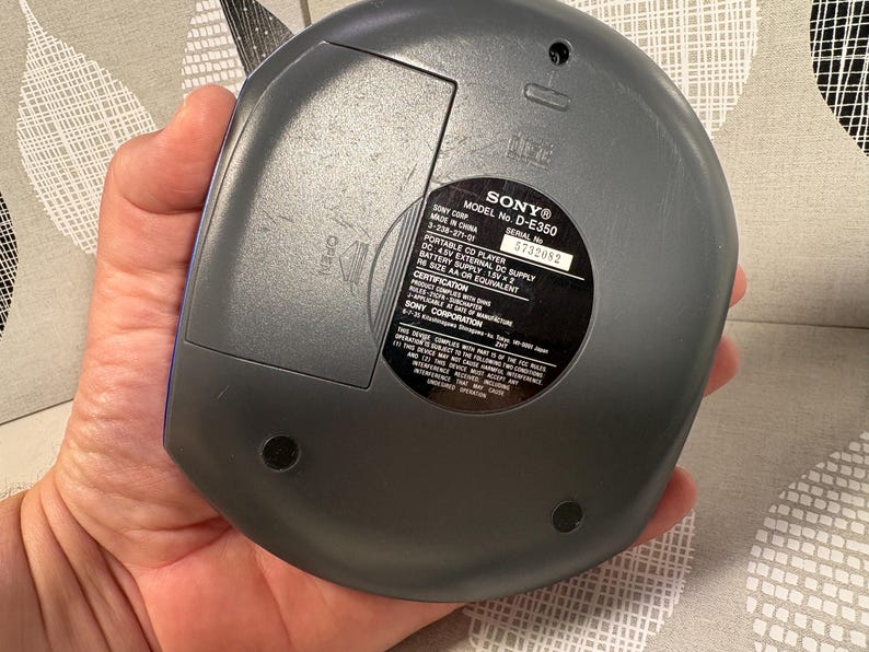 May include: A dark gray Sony portable CD player, model D-E350, featuring a circular design with a battery compartment door labeled OPEN. The back panel includes a black circular sticker with technical specifications, serial number 5732082, and regulatory text.