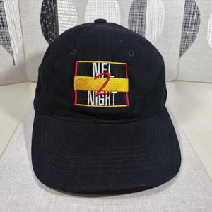 May include: Black baseball cap with a curved brim. The cap features an embroidered design with the text "NFL NIGHT" in white, red, and yellow. The cap is made of a soft fabric and has a rounded crown.