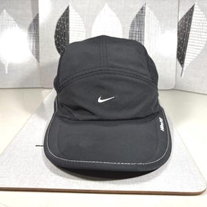 Może przedstawiać: Czarna czapka Nike z zakrzywionym daszkiem i białym logo Nike. Czapka ma strukturalny design i posiada napis "Nike-Fit" z boku. Leży na jasnej powierzchni.