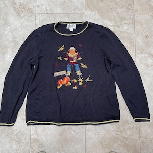 Könnte beinhalten: Ein dunkelblauer Langarm-Pullover mit gelbem Besatz an Ausschnitt und Ärmelbündchen. Der Pullover zeigt ein gesticktes Bild eines Vogelscheuchens mit Kürbissen und dem Text "Pumpkin Patch".