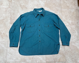 May include: A teal blue long-sleeved button-down shirt with two chest pockets and a single button cuff. The shirt has a white tag with the word "Wool" on it.