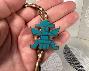Könnte beinhalten: Ein türkis-silbernes Bolo-Krawatte mit geometrischem Design. Der Anhänger ist eine stilisierte Figur mit türkisfarbener Einlage, vor einer geflochtenen Lederschnur in Braun- und Cremetönen. Ein Accessoire im Südwest-Stil.