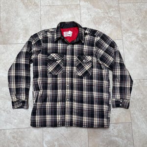 May include: Black, white, and tan plaid flannel shirt with a red quilted lining. The shirt has two chest pockets, a button-down front, and long sleeves. The collar and cuffs are black.
