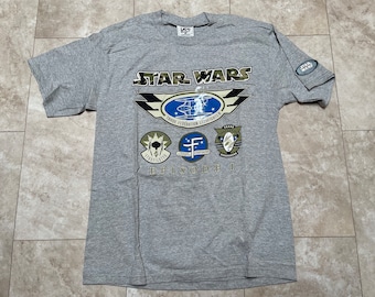 Peut inclure: Un t-shirt gris Star Wars Episode I avec un graphique d'un chasseur TIE et le texte "Trade Federation Starfighter". Le t-shirt présente également trois graphiques plus petits de différents véhicules Star Wars.