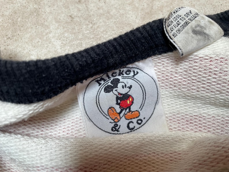 May include: White and pink striped sweatshirt with a black and white ringer neckline. The sweatshirt has a white circular patch with the text "Mickey & Co." and a cartoon image of Mickey Mouse.