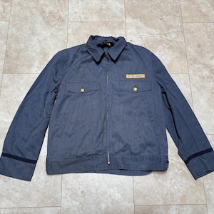 Op de afbeelding: Een blauw spijkerjack met een kraag, twee borstzakken en een ritssluiting. De jas heeft goudkleurige knopen en een rechthoekige patch met de tekst "LETTER CARRIER". Zwarte strepen sieren de mouwen.