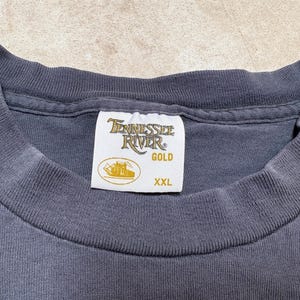May include: Close-up of a dark grey t-shirt collar with a white label. The label reads "Tennessee River GOLD" with a boat graphic and "XXL" size.