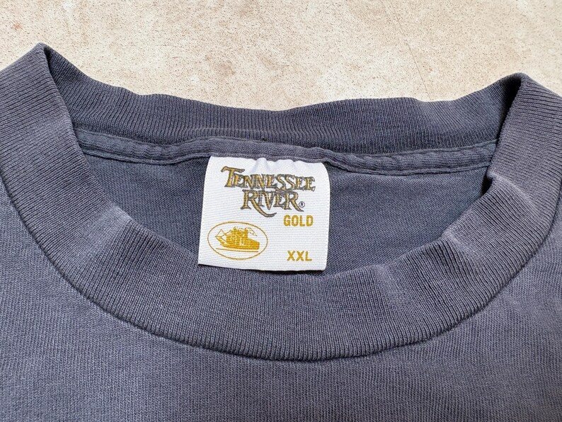 May include: Close-up of a dark grey t-shirt collar with a white label. The label reads "Tennessee River GOLD" with a boat graphic and "XXL" size.