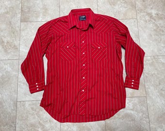 May include: A red and white striped long-sleeve button-up shirt with a pointed collar and two chest pockets. The shirt has a Wrangler label on the left side of the chest.