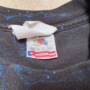 May include: Close-up of a black t-shirt with blue splatter paint design. The tag reads "ADULT FRESHRSHRUNK FRUIT OF THE LOOM MADE IN U.S.A."