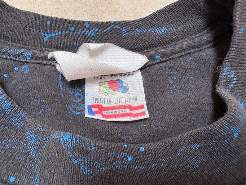 May include: Close-up of a black t-shirt with blue splatter paint design. The tag reads "ADULT FRESHRSHRUNK FRUIT OF THE LOOM MADE IN U.S.A."