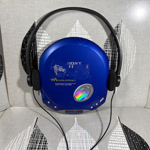 May include: A blue Sony Walkman portable CD player with black over-ear headphones. The circular device features the Sony logo, Walkman branding, and ESP MAX text on the front, with a small circular window showing a rainbow-colored disc inside.