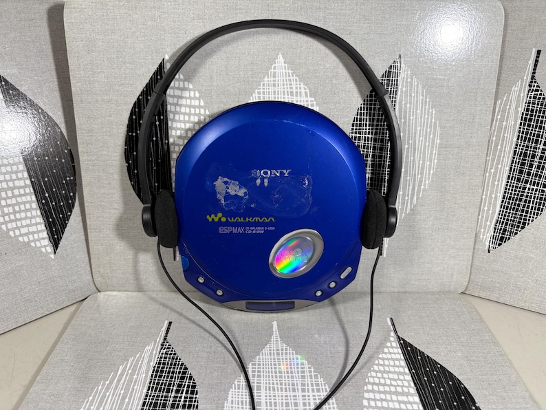May include: A blue Sony Walkman portable CD player with black over-ear headphones. The circular device features the Sony logo, Walkman branding, and ESP MAX text on the front, with a small circular window showing a rainbow-colored disc inside.