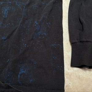 May include: Black long-sleeve shirt with a blue graphic design. The shirt features a ribbed cuff on the sleeve. The fabric appears to be a soft material. The design is visible on the front of the shirt.