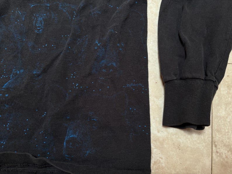 May include: Black long-sleeve shirt with a blue graphic design. The shirt features a ribbed cuff on the sleeve. The fabric appears to be a soft material. The design is visible on the front of the shirt.