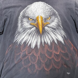 May include: A black t-shirt with a detailed illustration of a bald eagle's head. The eagle has white feathers on its head and neck, a yellow beak, and piercing yellow eyes. The chest feathers are brown.