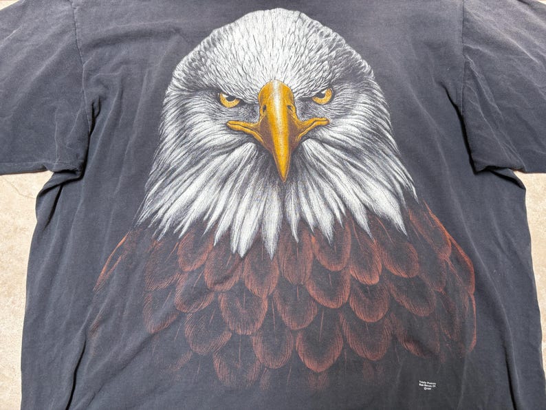 May include: A black t-shirt with a detailed illustration of a bald eagle's head. The eagle has white feathers on its head and neck, a yellow beak, and piercing yellow eyes. The chest feathers are brown.