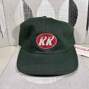 May include: A dark green baseball cap with a curved brim. The cap features a red oval patch with the letters "KK" in white, topped with crowns. The cap is a classic style, suitable for casual wear.
