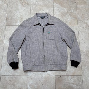 May include: A light gray zip-up jacket with a small chest pocket and black cuffs. The jacket has a collar and a small emblem on the chest. The jacket is made of a textured fabric.
