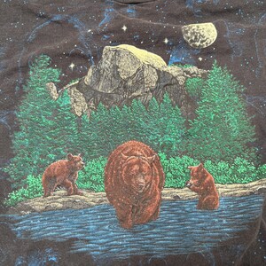May include: Black t-shirt featuring a nature scene with three bears in a river, a mountain, and evergreen trees. The background has a starry night sky with a moon. The bears are brown, and the trees are green.