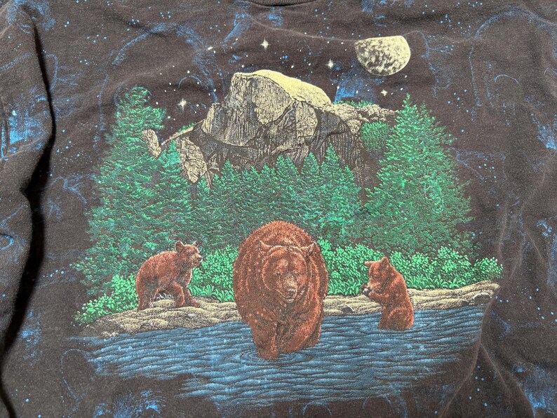 May include: Black t-shirt featuring a nature scene with three bears in a river, a mountain, and evergreen trees. The background has a starry night sky with a moon. The bears are brown, and the trees are green.