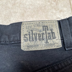 May include: Black denim jeans with a Levi Strauss & Co. SilverTab label. The label is brown with white text and a design of a pair of jeans.