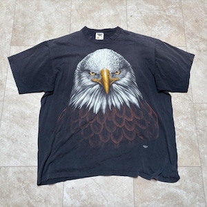 May include: A dark grey t-shirt with a detailed eagle head graphic. The eagle has white feathers, a yellow beak, and intense yellow eyes. The shirt is made of a soft material, likely vintage.