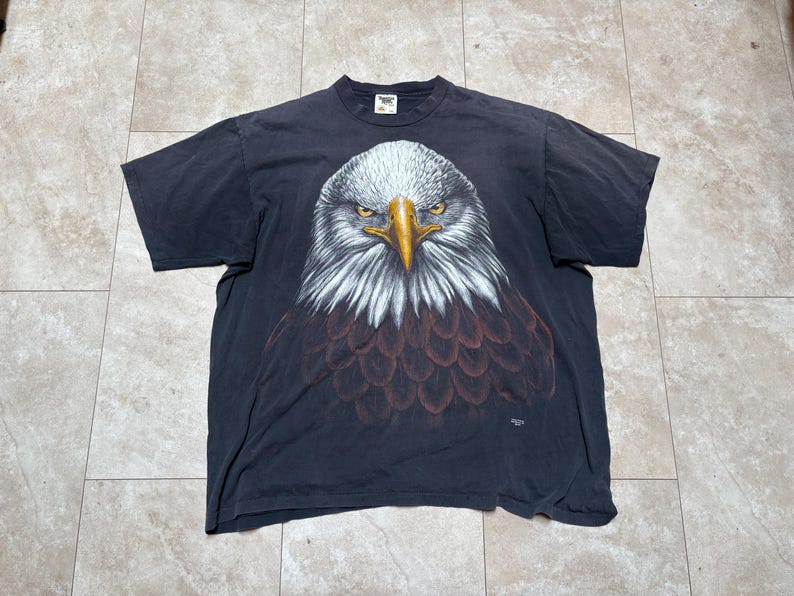 May include: A dark grey t-shirt with a detailed eagle head graphic. The eagle has white feathers, a yellow beak, and intense yellow eyes. The shirt is made of a soft material, likely vintage.