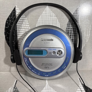 May include: A silver and blue Panasonic SL-SV570 portable CD player with an integrated FM/AM radio tuner and MP3 playback capability. The device features a digital display, D.SOUND anti-skip system, and is paired with black over-ear headphones.