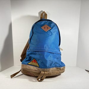 May include: Blue and tan backpack with a front pocket and shoulder straps. The backpack has a brown leather patch and a logo on the front. The backpack is made of nylon and canvas.