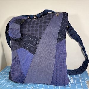 May include: A patchwork shoulder bag in shades of blue. The bag features various textured fabrics, including polka dots, stripes, and solid panels. It has a long, adjustable strap and metal grommets. The bag is approximately 10 inches wide and 12 inches tall.