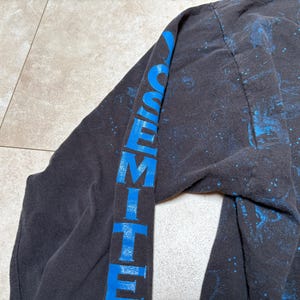 May include: Black long-sleeve shirt with the word "YOSEMITE" printed vertically in blue on the sleeve. The shirt has a nature-themed design with blue and green accents.