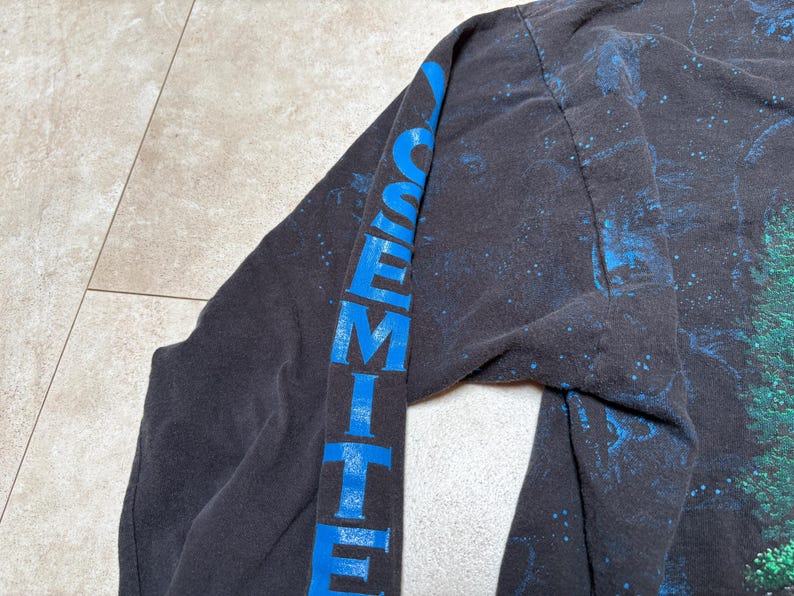 May include: Black long-sleeve shirt with the word "YOSEMITE" printed vertically in blue on the sleeve. The shirt has a nature-themed design with blue and green accents.