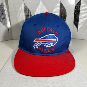 Puede incluir: Una gorra de béisbol azul y roja con las palabras "BUFFALO BILLS" bordadas en rojo y blanco. La gorra tiene una visera roja y una corona azul con el logotipo del equipo. Es un accesorio deportivo.